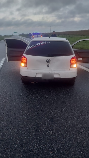 Automóvil blanco con puertas abiertas y baúl abollado sobre una ruta mojada frente a una patrulla policial y un agente