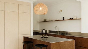 Vista de una cocina moderna con gabinetes de madera clara, una isla de madera oscura con encimera de acero inoxidable, y una lámpara colgante de mimbre.