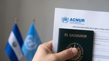 Mano adulta sostiene un pasaporte de El Salvador y una hoja de solicitud de asilo de ACNUR. Al fondo se observan las banderas de El Salvador y Naciones Unidas.