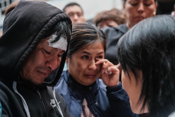 Un hombre con sudadera negra y vendaje en la frente mira hacia abajo, junto a una mujer que se toca el ojo. Varias personas al fondo
