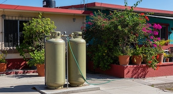 Vista exterior de dos tanques de gas LP verdes conectados por una manguera, frente a una casa mexicana con techo rojo, plantas frondosas y flores rosadas y rojas bajo el sol.