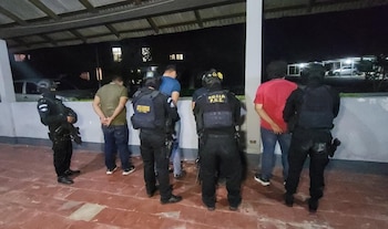 Cuatro agentes policiales uniformados y equipados con chalecos tácticos rodean a cinco hombres, tres de ellos con las manos esposadas, en un área techada de noche