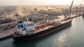 Un buque cisterna con la bandera de Argentina junto a un muelle con grúas, contenedores y la bandera de India al fondo.