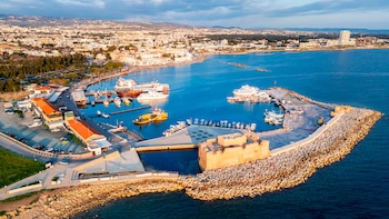 Vista aérea de un puerto con un castillo de piedra, numerosos barcos atracados, edificios en la costa, y una ciudad al fondo con montañas