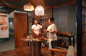 Dos mujeres conversan junto a una mesa redonda con objetos de madera bajo dos lámparas colgantes en un puesto de exhibición