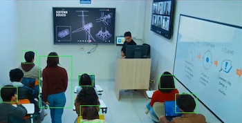 Salón de clases con un profesor al frente, varios estudiantes en escritorios, pantallas mostrando diagramas y videollamadas, y un pizarrón