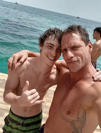 Dos hombres, uno mayor y uno joven, posan sonrientes y mojados junto a un muelle de madera, con el mar y un barco de fondo