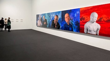 En una galería de arte, tres personas miran una larga serie de paneles que muestran retratos distorsionados y una figura similar a un busto, con paredes blancas y suelo oscuro