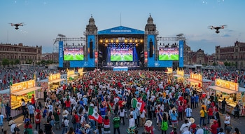 Multitud en el Zócalo de CDMX viendo fútbol en pantallas gigantes del FIFA Fan Festival; puestos de comida, edificios históricos y dos drones visibles en el cielo.
