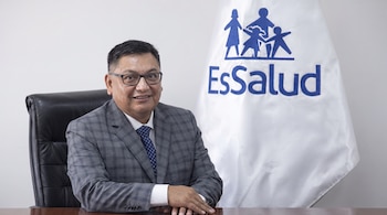 Hombre con traje y gafas sentado en un escritorio de madera, con bandera blanca de EsSalud con logo azul de familia detrás.