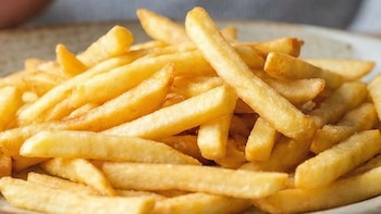 Primer plano de un plato con papas fritas en una mesa de madera. Al fondo, una silueta borrosa de una panza de embarazada y una mano en posición de rechazo.