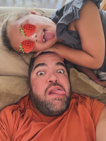Darío Barassi y una niña rubia con fresas en los ojos, ambos acostados y sacando la lengua con expresiones graciosas