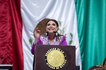 Clara Brugada viste un saco morado y habla en un podio con el escudo del Congreso de la Ciudad de México y una bandera de fondo