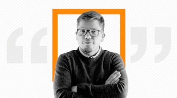 Retrato en escala de grises de un hombre con gafas y barba, brazos cruzados, enmarcado por un cuadrado naranja sobre fondo blanco con líneas y comillas.