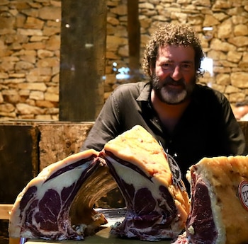 Hombre barbudo de cabello rizado con camisa oscura sonríe detrás de tres grandes cortes de carne roja con hueso y grasa visible