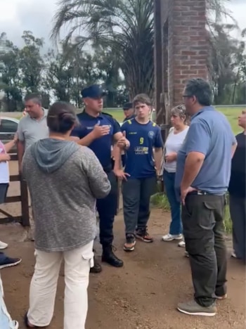 Un policía uniformado habla con varias personas frente a un pilar de ladrillo y un portón en un camino de tierra con árboles al fondo