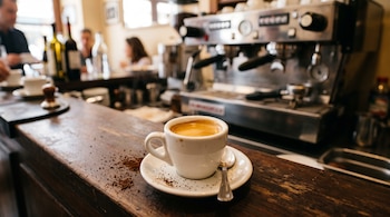 Taza blanca de café espresso en un plato con cuchara sobre una barra de madera con café molido y una máquina metálica al fondo.