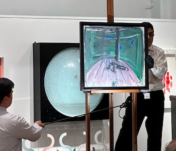 Hombre con guante negro sostiene una pintura abstracta titulada 'TRES NOCHES' sobre un atril. Otro hombre visible, y obra de arte circular en el fondo