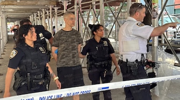 Cuatro agentes de policía con uniformes y equipo táctico acompañan a un hombre esposado bajo andamios. Se observa una cinta "Police Line Do Not Cross".