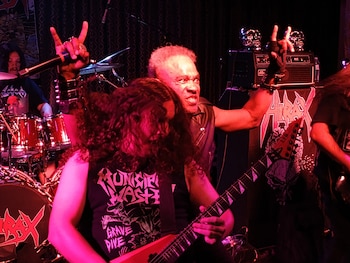 Un vocalista con chaleco de cuero grita y hace un gesto de rock, junto a un guitarrista de pelo rizado tocando una guitarra roja en un concierto con luces rojas