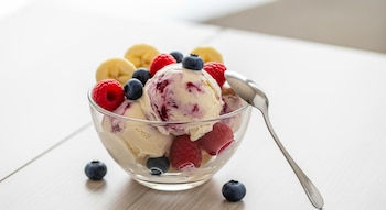 Cuenco de vidrio con varias bolas de helado, rodajas de plátano, frambuesas y arándanos frescos, con una cuchara de metal a la derecha.