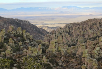 Formaciones y pináculos de roca erosionada en un cañón con vegetación dispersa, un valle árido al fondo y montañas bajo el cielo claro