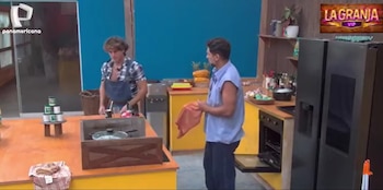 Dos hombres en una cocina. Uno lleva delantal y camisa a cuadros, el otro sostiene un paño cerca de un horno abierto y un refrigerador