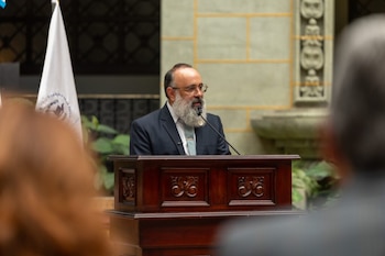 Un hombre con barba y gafas habla en un micrófono. Se encuentra detrás de un podio de madera. Una bandera blanca con un emblema se ve a la izquierda. Hay personas en primer plano