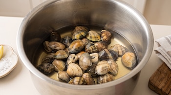 Almejas frescas sumergidas en agua dentro de una olla de acero inoxidable, junto a un plato con sal, un trozo de limón y un paño.