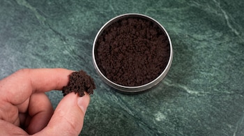 Mano sosteniendo una porción de tabaco húmedo oscuro junto a una lata metálica abierta sobre una superficie de mármol verde.