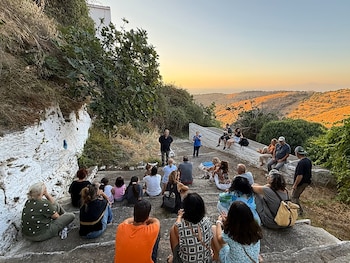 Personas sentadas en escalones de piedra al aire libre mientras dos personas de pie hablan frente a ellas con colinas al fondo
