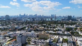 Vista aérea diurna de un denso paisaje urbano con edificios de varios pisos, rascacielos al fondo, un cuerpo de agua azul y nubes blancas en el cielo