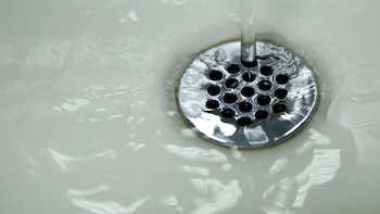 Primer plano de agua transparente fluyendo de un grifo hacia un desagüe circular cromado con orificios negros en un lavabo blanco