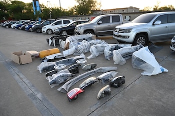 Primer plano de parachoques, faros, llantas y otras autopartes envueltas en plástico en el suelo. Al fondo, una fila de vehículos estacionados, incluyendo SUV y coches