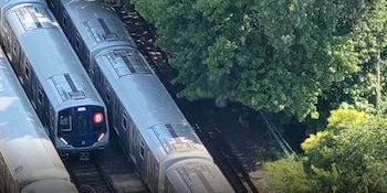 Vista aérea de varios trenes grises en vías férreas paralelas. Uno de los trenes exhibe la letra B en un círculo rojo en su frente, junto a árboles