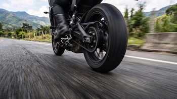Rueda trasera y parte inferior de una motocicleta en movimiento sobre una carretera de asfalto con montañas al fondo.