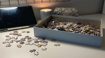 Mesa blanca con caja de rompecabezas, piezas esparcidas, tableta negra con lápiz digital, vela encendida y estuche floral en un ambiente tenue.