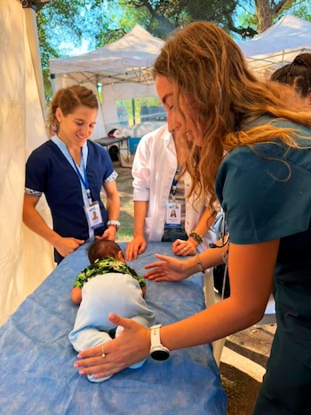 Tres médicas, incluida Paula Pareto, examinan a un bebé boca abajo en una camilla azul. La escena ocurre en un campamento médico al aire libre bajo carpas