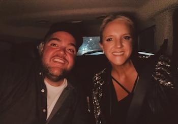 Primer plano de un hombre barbudo y una mujer rubia, ambos sonriendo ampliamente, sentados dentro de un coche oscuro por la noche con luces borrosas al fondo