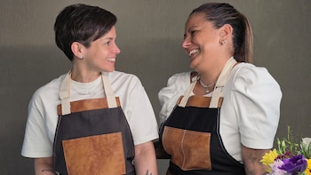 Dos mujeres vestidas con camisetas blancas y delantales oscuros se miran y sonríen, con un ramo de flores a la derecha
