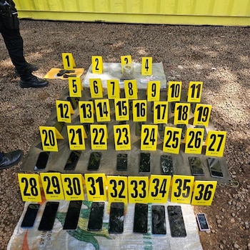 Arma, bolsa con sustancia, pastillas, 29 teléfonos. Objetos con marcadores de evidencia amarillos (1-36). Sobre lona en suelo de grava. Piernas