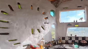 Pared de adobe con botellas de vidrio incrustadas en abanico, junto a ventanas rectangulares y una mesada de cocina con pileta