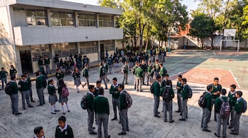 Estudiantes con suéteres verdes y pantalones grises reunidos en un patio escolar junto a un edificio y una cancha de baloncesto.