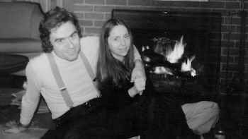 Fotografía en blanco y negro de Ted Bundy y Elizabeth Kendall sentados en el suelo frente a una chimenea de ladrillo con fuego, él la abraza