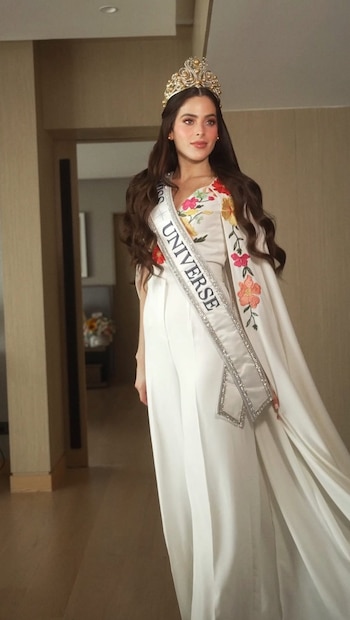 Mujer de cabello castaño y largo con corona dorada y banda "MISS UNIVERSE" posa en interior. Viste un traje blanco con bordados florales de colores y una capa