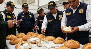 Agentes de seguridad con uniformes azules y gorras negras inspeccionan una mesa llena de panes redondos. Varios panes están abiertos mostrando paquetes blancos en su interior.