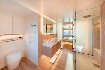 Baño con inodoro blanco, lavabo sobre mueble de madera, espejo iluminado, ducha con mampara de vidrio y acceso a un dormitorio