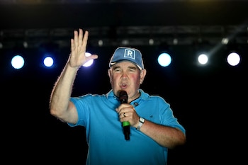 Primer plano de Rafael López Aliaga en un escenario oscuro, con gorra azul y polo del mismo color, hablando por un micrófono y levantando la mano derecha