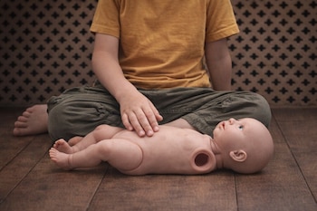 Un niño con camiseta amarilla y pantalones verdes se sienta en el suelo junto a un muñeco de bebé sin un brazo. La mano del niño toca el abdomen del muñeco.