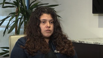 Una mujer de cabello largo y ondulado viste una chaqueta azul oscura con un micrófono de solapa en una sala interior